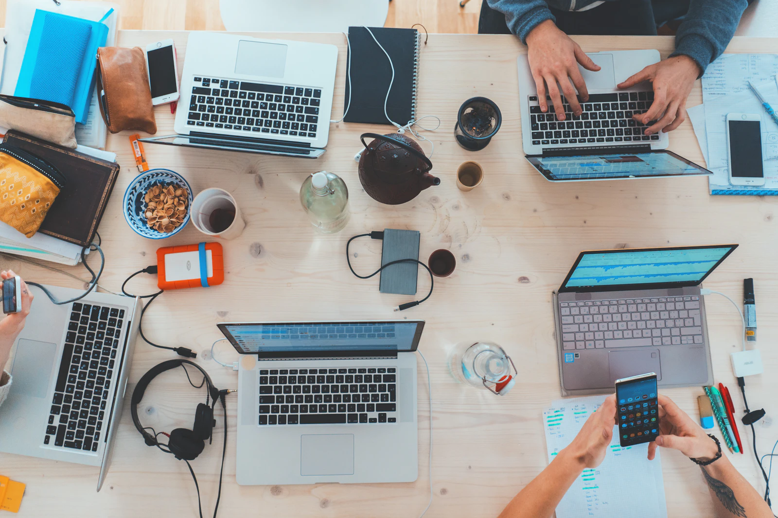 A desk crowded with laptops, phones, notebooks and cables seen from above
